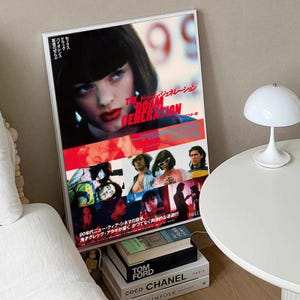 May include: A framed movie poster for "The Doom Generation" with a woman's face in the top section. The poster has a red and white colour scheme with Japanese text. The poster is propped up against a wall, with books and a lamp nearby.