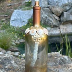 May include: A gold-toned glass bottle oil lamp featuring a white wick extending from a brass-colored collar. The neck is decorated with a ring of small white seashells and amber-colored beads, with a cross emblem embossed on the front surface.