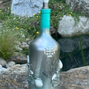 May include: A silver-painted glass bottle with a bright turquoise neck and base. The bottle features a raised Jerusalem cross emblem on the front and is decorated with various white and clear glass pieces attached to the lower section, topped with a wick.