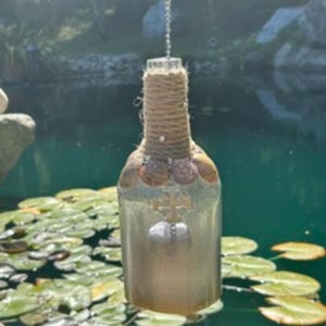 May include: A hanging glass bottle ornament featuring a neck wrapped in tan jute twine, decorated with a row of small seashells and a gold-toned cross charm. The bottle has a frosted, semi-translucent finish and hangs from a silver metal chain.
