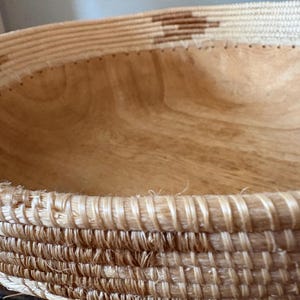 Op de afbeelding: Een geweven mand met een lichtbruin houten interieur. De rand van de mand is gemaakt van dicht geweven natuurlijke vezels, met een decoratief patroon van donkerbruine vormen. De houtnerf aan de binnenkant is zichtbaar.