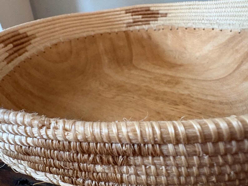 Op de afbeelding: Een geweven mand met een lichtbruin houten interieur. De rand van de mand is gemaakt van dicht geweven natuurlijke vezels, met een decoratief patroon van donkerbruine vormen. De houtnerf aan de binnenkant is zichtbaar.