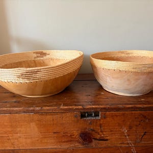 Op de afbeelding: Twee houten kommen met geweven randen. De kommen zijn lichtbruin met een natuurlijke houtnerf. De randen zijn gemaakt van geweven strokleurig materiaal. De kommen staan op een houten oppervlak.
