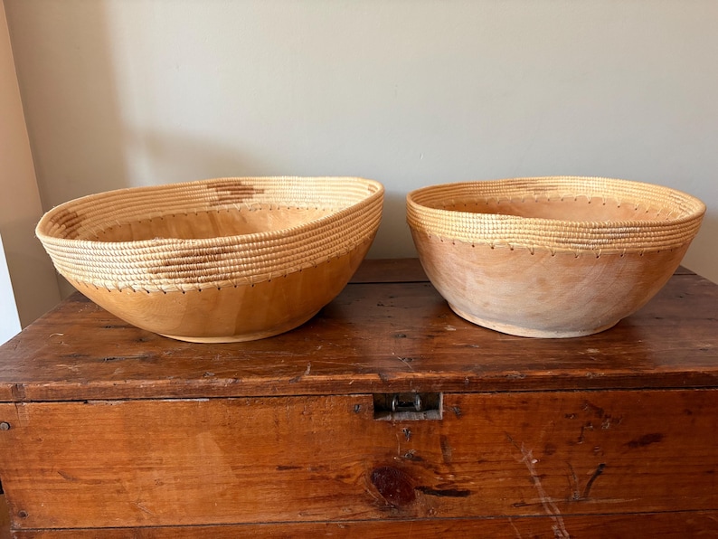 Op de afbeelding: Twee houten kommen met geweven randen. De kommen zijn lichtbruin met een natuurlijke houtnerf. De randen zijn gemaakt van geweven strokleurig materiaal. De kommen staan op een houten oppervlak.