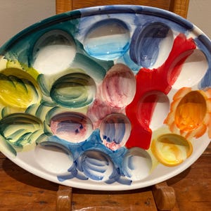 May include: Oval ceramic serving platter with a colorful, abstract design. The platter features multiple indentations, each painted with a different color, including blue, red, yellow, and green. The rim is white, and the overall style is artistic and decorative.