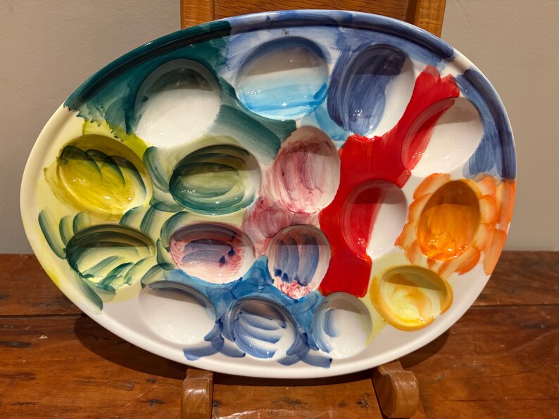May include: Oval ceramic serving platter with a colorful, abstract design. The platter features multiple indentations, each painted with a different color, including blue, red, yellow, and green. The rim is white, and the overall style is artistic and decorative.