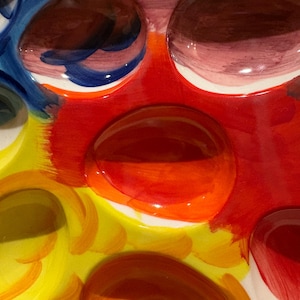 Pu&ograve; includere: Primo piano di un colorato portauova in ceramica. Il portauova presenta un design vibrante con tonalit&agrave; rosse, gialle, arancioni, blu e marroni. Il portauova ha pi&ugrave; rientranze circolari per contenere le uova.