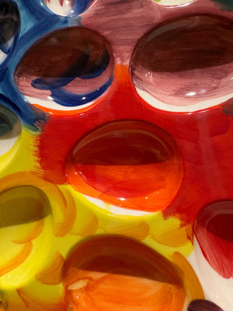 Pu&ograve; includere: Primo piano di un colorato portauova in ceramica. Il portauova presenta un design vibrante con tonalit&agrave; rosse, gialle, arancioni, blu e marroni. Il portauova ha pi&ugrave; rientranze circolari per contenere le uova.