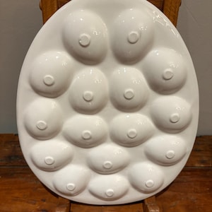 Pu&ograve; includere: Piatto da portata in ceramica bianca con un design unico caratterizzato da molteplici rientranze arrotondate a forma di capezzolo. Il piatto &egrave; di forma ovale e poggia su una sedia di legno. La finitura lucida e la forma insolita lo rendono un pezzo decorativo distintivo.