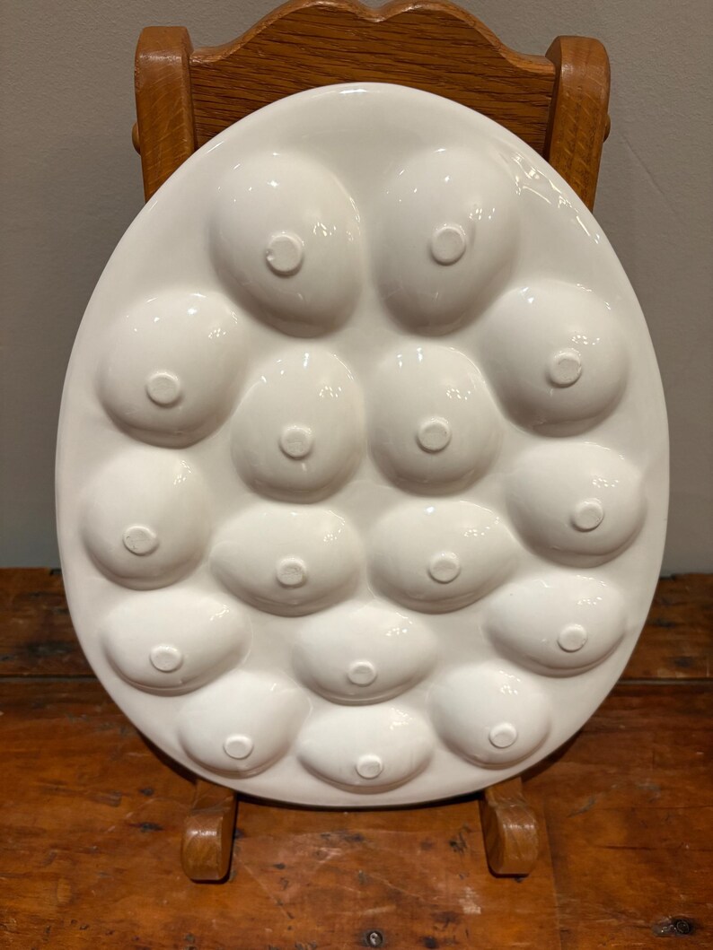 Pu&ograve; includere: Piatto da portata in ceramica bianca con un design unico caratterizzato da molteplici rientranze arrotondate a forma di capezzolo. Il piatto &egrave; di forma ovale e poggia su una sedia di legno. La finitura lucida e la forma insolita lo rendono un pezzo decorativo distintivo.