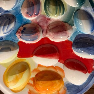 May include: A colorful, oval-shaped ceramic serving platter with multiple egg-shaped indentations. The platter features a vibrant palette of colors, including blue, red, yellow, and green, with a glossy finish. Ideal for serving deviled eggs or appetizers.