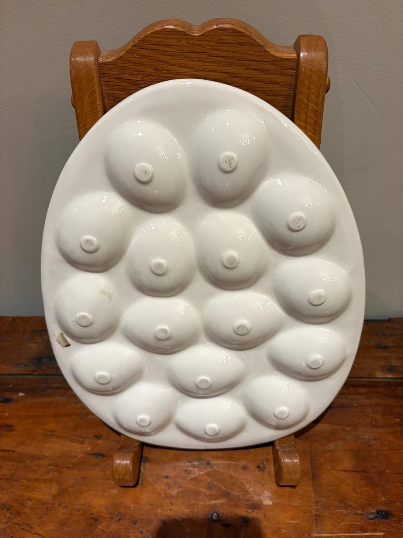 May include: White ceramic egg serving platter with 15 egg-shaped indentations. The platter is oval and has a glossy finish. The platter is sitting on a wooden surface.
