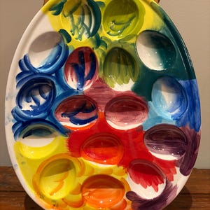Pu&ograve; includere: Un vassoio da portata in ceramica a forma di uovo con 12 rientranze a forma di uovo. Il vassoio &egrave; dipinto con una vibrante gamma di colori, tra cui giallo, blu, verde, rosso e arancione. Il vassoio &egrave; supportato da un supporto in legno.