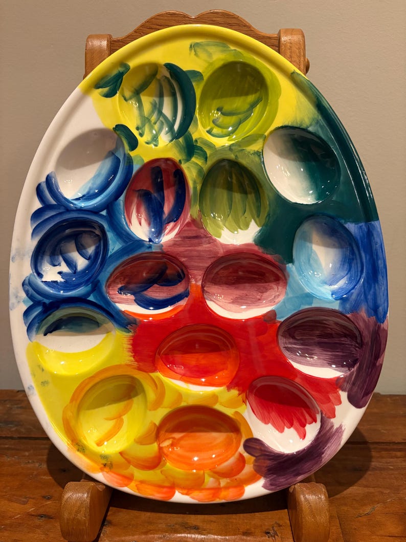 Pu&ograve; includere: Un vassoio da portata in ceramica a forma di uovo con 12 rientranze a forma di uovo. Il vassoio &egrave; dipinto con una vibrante gamma di colori, tra cui giallo, blu, verde, rosso e arancione. Il vassoio &egrave; supportato da un supporto in legno.