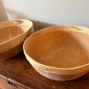 Op de afbeelding: Twee houten kommen met geweven randen. De kommen zijn lichtbruin met een natuurlijke houtnerf. De randen zijn lichtbeige met een geweven patroon. De kommen staan op een donkerbruin houten oppervlak.