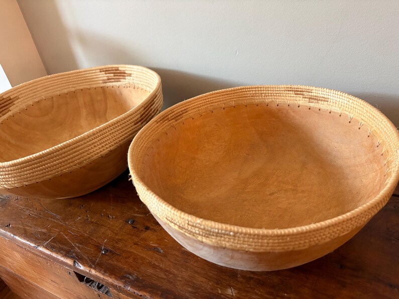 Op de afbeelding: Twee houten kommen met geweven randen. De kommen zijn lichtbruin met een natuurlijke houtnerf. De randen zijn lichtbeige met een geweven patroon. De kommen staan op een donkerbruin houten oppervlak.