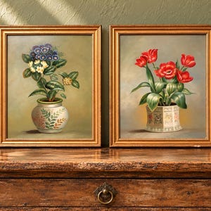 May include: Two framed oil paintings in gold-toned frames. The left painting depicts blue and white flowers in a patterned ceramic vase. The right painting shows red tulips in a hexagonal, floral-patterned ceramic pot against a soft background.