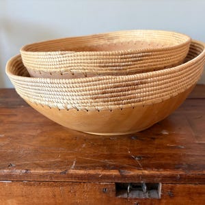 Op de afbeelding: Twee geneste decoratieve kommen. De onderste kom is lichtbruin hout met een geweven rand. De bovenste kom is kleiner, met een vergelijkbare geweven rand. De kommen staan op een houten oppervlak.