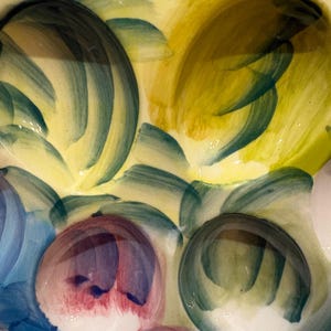 May include: Close-up of a ceramic plate with a hand-painted design. The plate features a yellow background with brushstrokes of green, blue, and red. The design includes rounded shapes and abstract patterns.