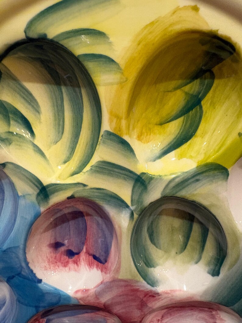 May include: Close-up of a ceramic plate with a hand-painted design. The plate features a yellow background with brushstrokes of green, blue, and red. The design includes rounded shapes and abstract patterns.