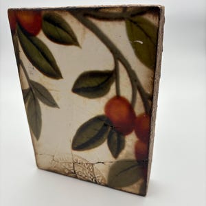 May include: A rectangular ceramic tile featuring a botanical design. The artwork depicts orange-red fruit and green leaves on a branch against a cream-colored background. The tile has a distressed, aged appearance with visible cracks.
