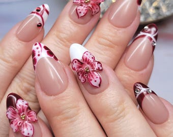 Può includere: Una mano con unghie lunghe a forma di mandorla. Le unghie presentano una base nude con vari disegni: punte bianche con pois rossi, punte bordeaux e accenti floreali rosa 3D. Alcune unghie hanno piccoli abbellimenti di gemme.