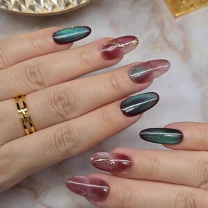 May include: Oval-shaped nails with a colour-shifting effect, transitioning from deep burgundy to emerald green. Some nails feature delicate gold wire accents. A gold ring is visible on one finger.