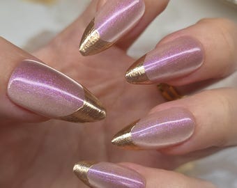 May include: Close-up of a hand with five almond-shaped nails. The nails have a pearlescent pink hue with a gold metallic French tip. The nails are smooth and shiny, reflecting light.