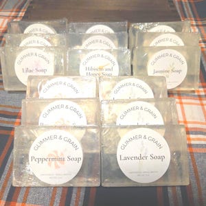 May include: Clear soap bars, individually wrapped, are stacked. Each bar has a label reading "Glimmer & Grain" with scents including Lilac, Hibiscus and Honey, Jasmine, Peppermint, and Lavender. The soaps are presented on a plaid surface.