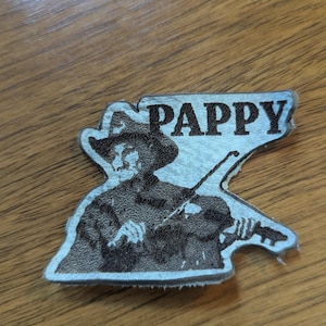 May include: A silver-colored, die-cut emblem featuring a fiddler playing a violin, with the word "PAPPY" above. The fiddler is wearing a hat. The emblem is on a wooden surface.