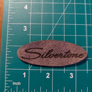 May include: A brown, oval-shaped leather patch with the word "Silvertone" embossed on it. The patch measures approximately 3 inches long. The patch is on a green cutting mat with inch measurements.