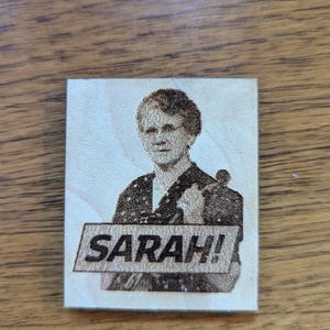 May include: A wooden plaque featuring a portrait of a woman with the name "SARAH!" etched below. The image is a detailed, grayscale engraving on a light-colored wood surface, showcasing the woman's likeness and the bold text.