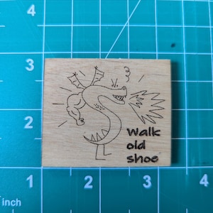 May include: A small, square wooden block with a laser-etched design of a cartoon dragon breathing fire. The text "walk old shoe" is below the dragon. The block measures approximately 2 inches by 2 inches.