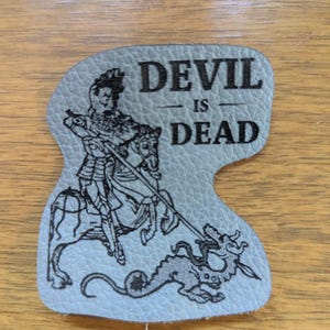 May include: A light blue, leather-like patch with the words "DEVIL IS DEAD" in black block letters. An illustration of a knight on horseback slaying a dragon is below the text. The patch has a unique, irregular shape.
