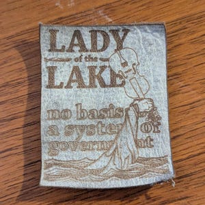 May include: A rectangular, light gray leather patch with the words "LADY of the LAKE" and "no basis a system of government" etched in brown. A violin is depicted above a figure in water.