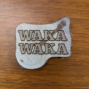 May include: A light-colored leather patch with the words "WAKA WAKA" in a brown, textured font. A brown, stylized design is above the text. The patch is on a wooden surface.