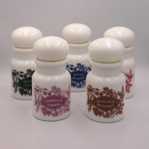 May include: Five white spice jars with rounded lids. Each jar has a different label with text and botanical illustrations in various colours, including green, purple, blue, and brown. The jars are arranged on a white surface.