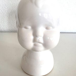 Könnte beinhalten: Eine weiße Keramik-Babykopfvase mit geschlossenen Augen und einem abgerundeten Kinn. Die Vase hat eine glatte, glänzende Oberfläche und einen Sockel. Das Design ist im Vintage- oder Antik-Stil gehalten und eignet sich für Blumengestecke oder als dekoratives Element.