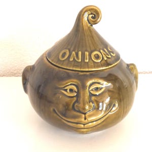 Może przedstawiać: Kapryśny, oliwkowozielony ceramiczny pojemnik na cebulę z uśmiechniętą twarzą i kręconym wierzchołkiem. Słowo "ONIONS" jest wytłoczone na pokrywce. Pojemnik ma pękaty kształt z małymi uchwytami w kształcie uszu po bokach.