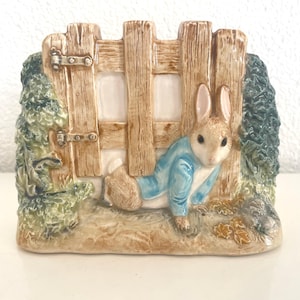 Może przedstawiać: Ceramiczna figurka przedstawiająca królika w niebieskiej kurtce wychodzącego z drewnianego płotu. Figurka ma szczegółowy, ręcznie malowany wzór z naturalnymi kolorami, w tym odcieniami brązu, zieleni i beżu. Scena przywodzi na myśl bajkę.