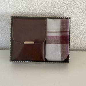 Può includere: Una scatola regalo rettangolare contenente un portafoglio marrone scuro con chiusura in metallo argentato e un fazzoletto bianco piegato con motivo a quadri bordeaux e grigio. Gli articoli sono presentati affiancati in una custodia trasparente.