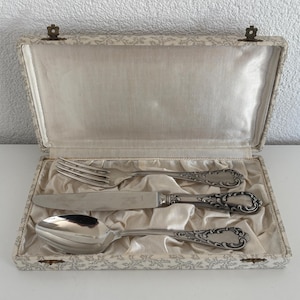May include: A silver-toned three-piece cutlery set comprising a fork, a knife, and a spoon, each featuring ornate scrollwork on the handles. The utensils are arranged horizontally inside a rectangular, cream-coloured fabric-lined presentation box.