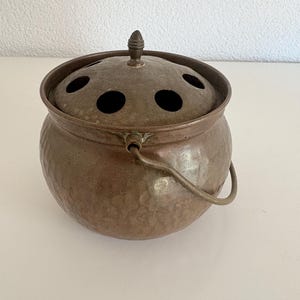 May include: A vintage bronze-colored metal incense burner with a round, hammered texture. The burner has a lid with several circular holes and a small handle. A curved handle is attached to the side of the pot.
