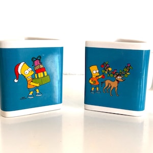 May include: Two square ceramic mugs with white handles and rims. The mugs have a teal blue background with cartoon images of Bart Simpson. One mug shows Bart in a Santa hat carrying gifts. The other shows Bart with a dog decorated with Christmas ornaments.