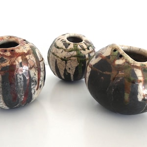 Op de afbeelding: Drie keramische vaten met een ronde vorm en een textuur. De vaten hebben een donker kleurenpalet met zwart, bruin en wit. Elk vat heeft een cirkelvormige opening aan de bovenkant en kleine gaatjes rond de rand.