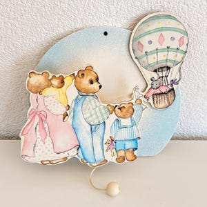 以下が含まれることがあります： 熱気球に乗った3匹のクマとネズミが描かれた木製の壁掛けプレート。クマはパステルカラーの服を着ており、下部には紐で小さな木製のビーズがぶら下がっています。背景は水色の円形になっています。