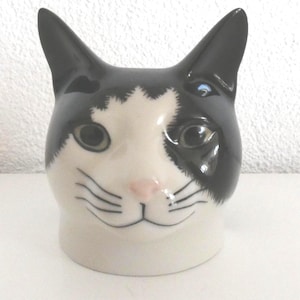 Könnte beinhalten: Eine keramische katzenförmige Tasse mit einem schwarz-weißen Design. Die Tasse hat ein weißes Gesicht mit schwarzen Markierungen um Augen und Ohren, eine rosa Nase und schwarze Schnurrhaare. Ein Neuheitenartikel.