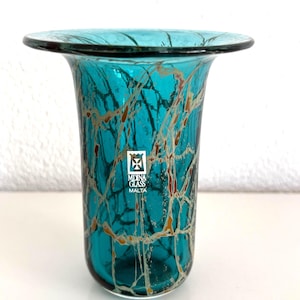 May include: A teal glass vase with a flared rim and a webbed pattern of white and amber lines throughout the body. A white rectangular sticker on the front displays a cross emblem above the text MDINA GLASS MALTA in black lettering.