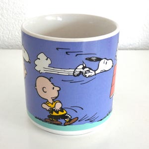 May include: A ceramic mug featuring a cartoon scene with Charlie Brown and Snoopy. The mug has a blue background with white clouds and illustrations of the characters. The mug has a white interior and a turquoise and white base.