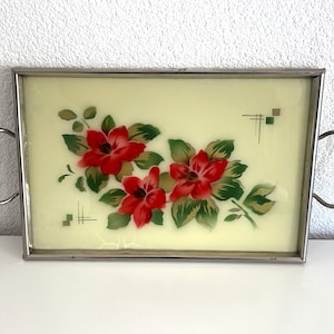 Könnte beinhalten: Rechteckiges Serviertablett mit hellgelbem Hintergrund und einem Blumendesign mit roten Blüten und grünen Blättern. Das Tablett hat einen silbernen Metallrahmen und Griffe, geeignet zum Servieren von Speisen oder zum Ausstellen von Dekorationsartikeln.